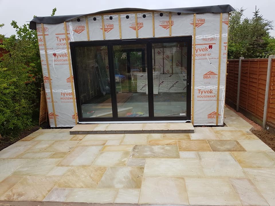 A small building under construction with a sliding glass door opening, surrounded by a partially finished stone patio and a wooden fence on one side.