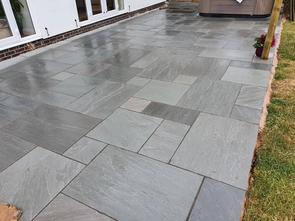 A newly paved outdoor patio area with rectangular gray stone tiles, adjoining a white house with large windows and a small garden with flowers.