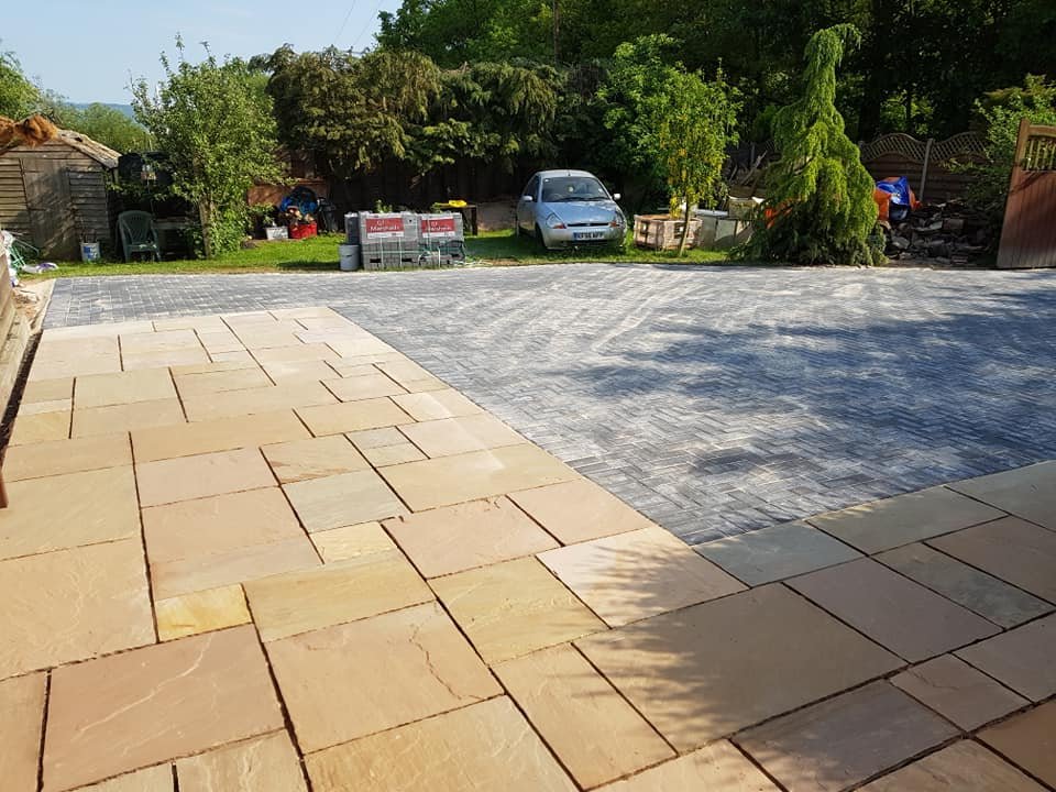 A backyard with a partially renovated patio featuring beige stone tiles and gray interlocking pavers, surrounded by green trees and garden furniture.