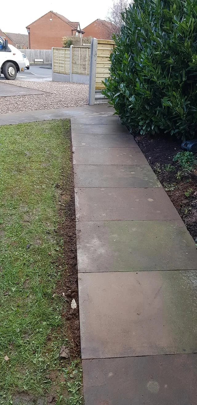 A sidewalk with concrete slabs next to a grass lawn and tall green bushes. There is a wooden gate and fence at the end of the sidewalk, with a parked white van and a residential street in the background.