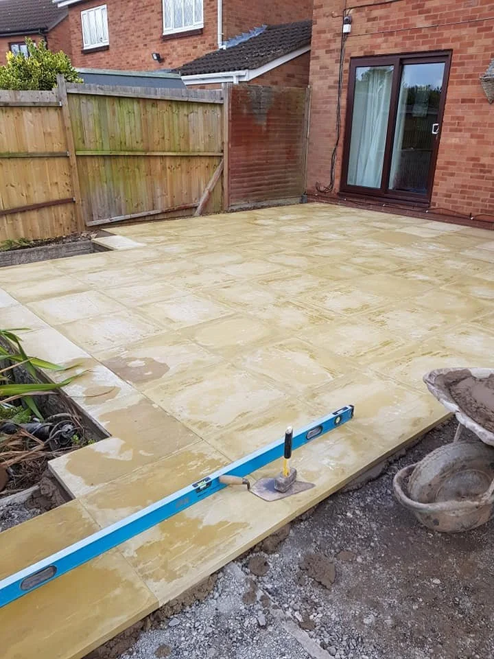 A backyard patio with newly installed yellowish stone tiles, ungrouted, next to a house with a sliding glass door; construction tools and materials visible, including a level, a trowel, and a large stone bowl.