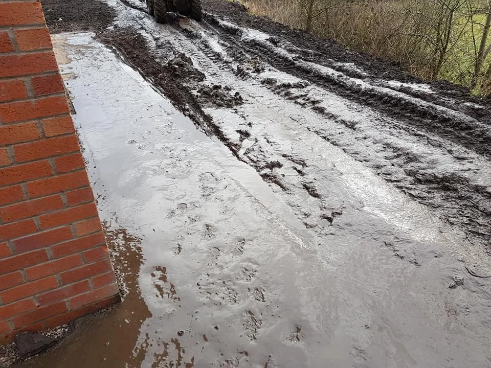 A muddy construction site with a wet, muddy ground, tire tracks, and footprints near a brick building wall.
