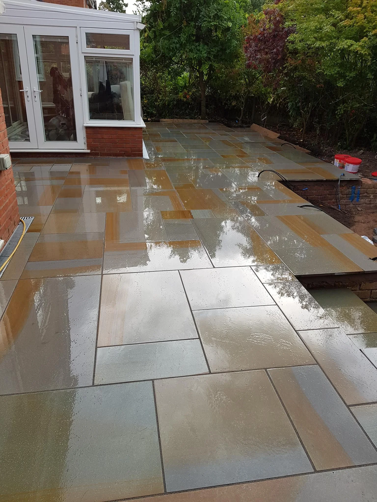 Wet brick patio extending from house with a conservatory, surrounded by green bushes and trees.