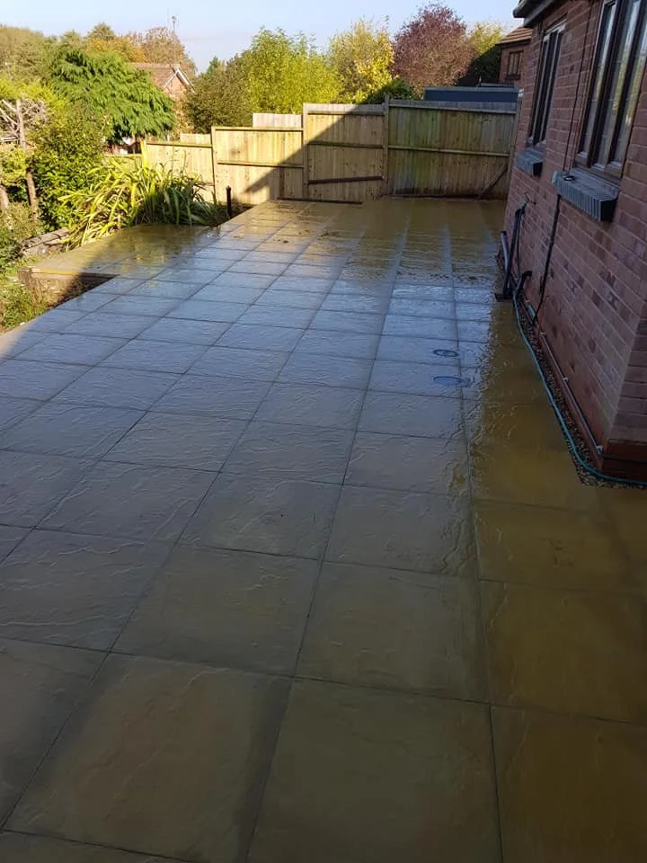Newly cleaned and wet outdoor patio with large square tiles, adjacent to a brick house with windows, surrounded by a wooden fence and trees in the background.
