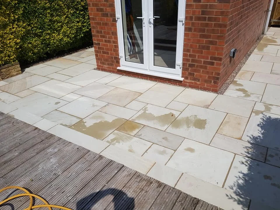 A backyard patio with newly installed light-colored stone tiles, partially wet, adjacent to a brick building with white-framed glass doors.