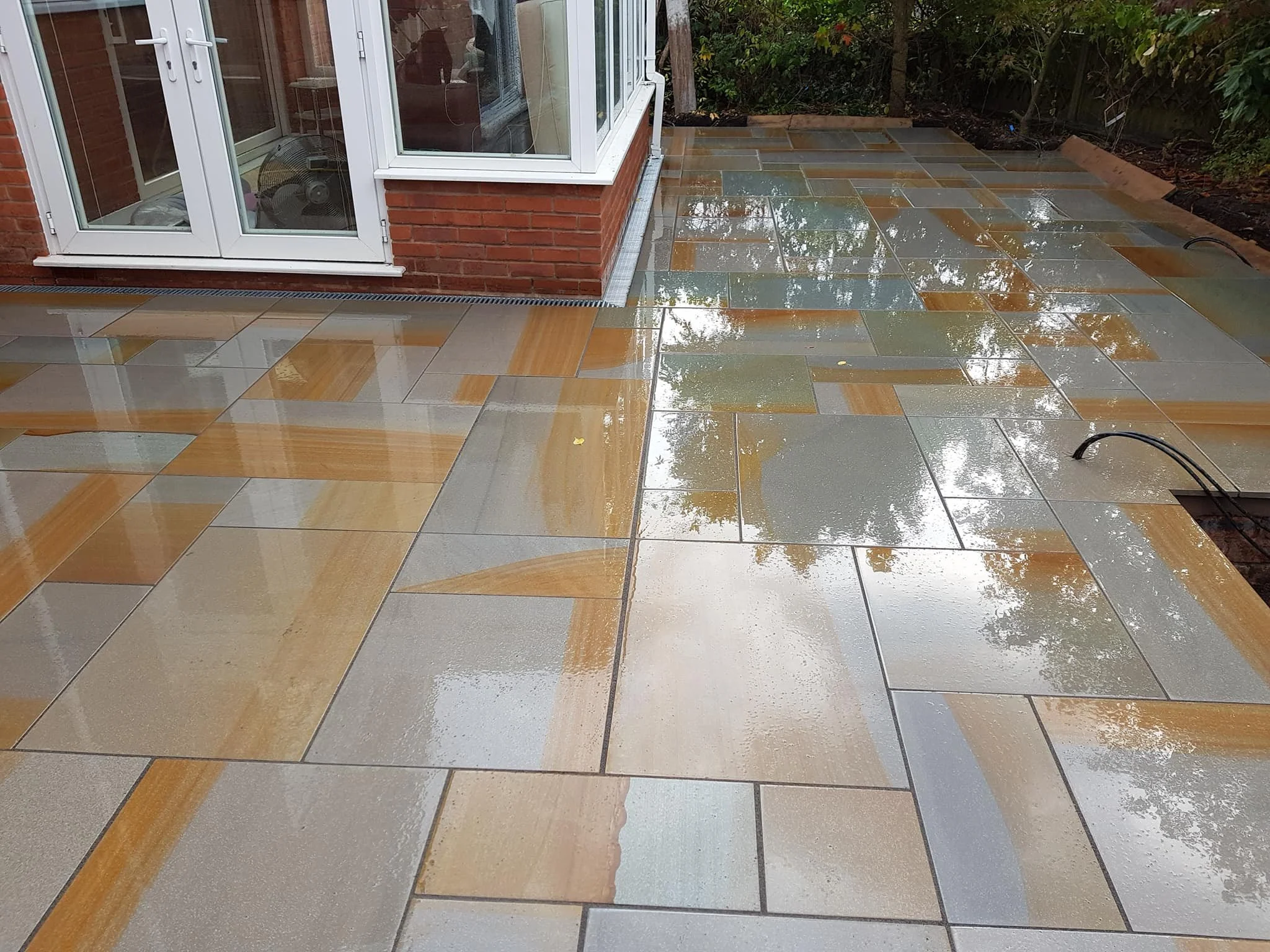 Rainy day showing freshly cleaned and wet stone patio outside a brick house with glass door and garden bushes in the background.