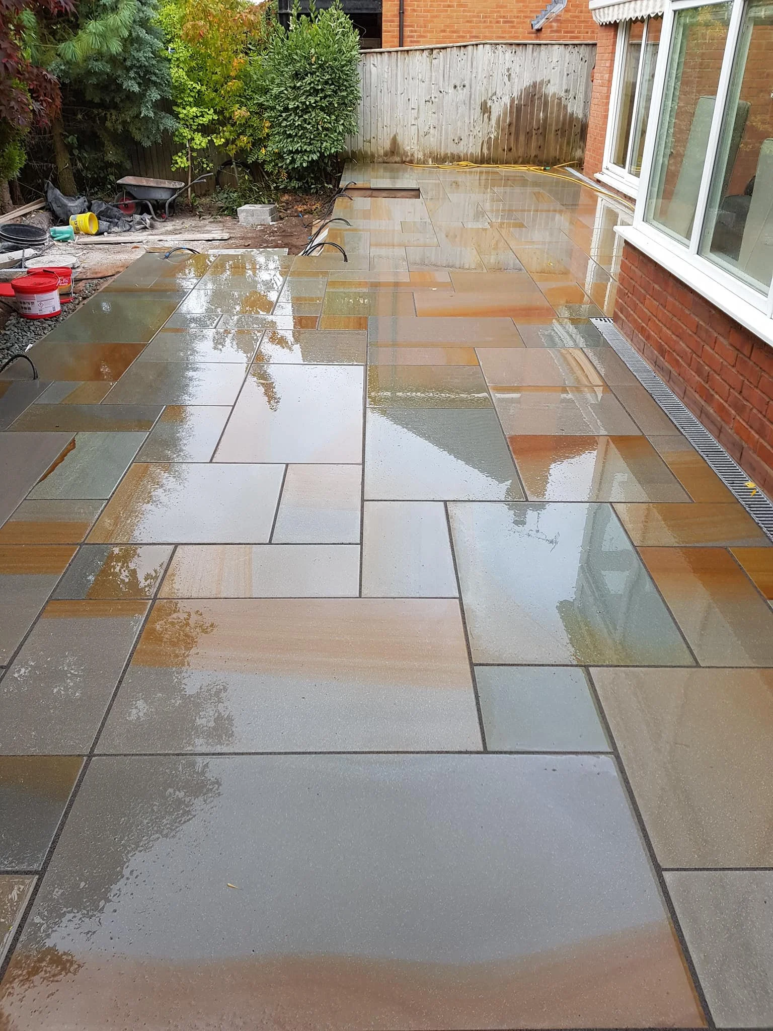 Recently finished wet patio with multicolored rectangular stone tiles in shades of tan, gray, and rust, with reflections of trees and sky on the surface, next to a brick house with large window.