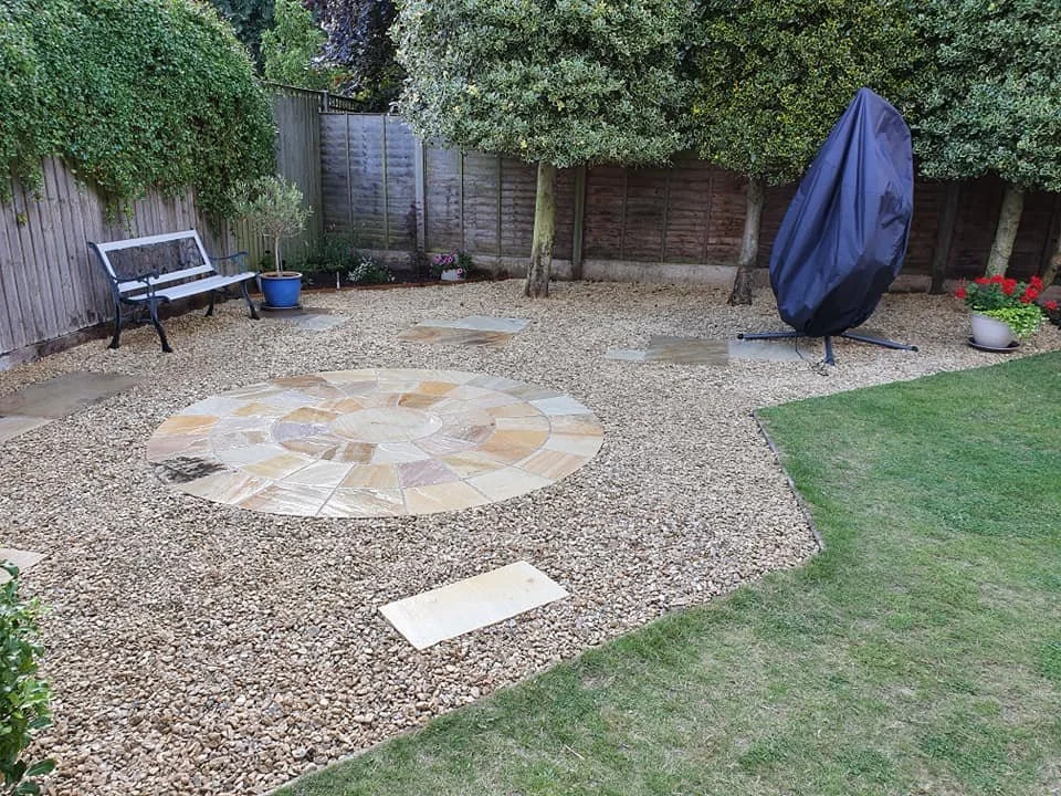 A backyard garden with a brick and gravel patio, a bench, potted plants, a covered outdoor heater, three trees, and a wooden fence.