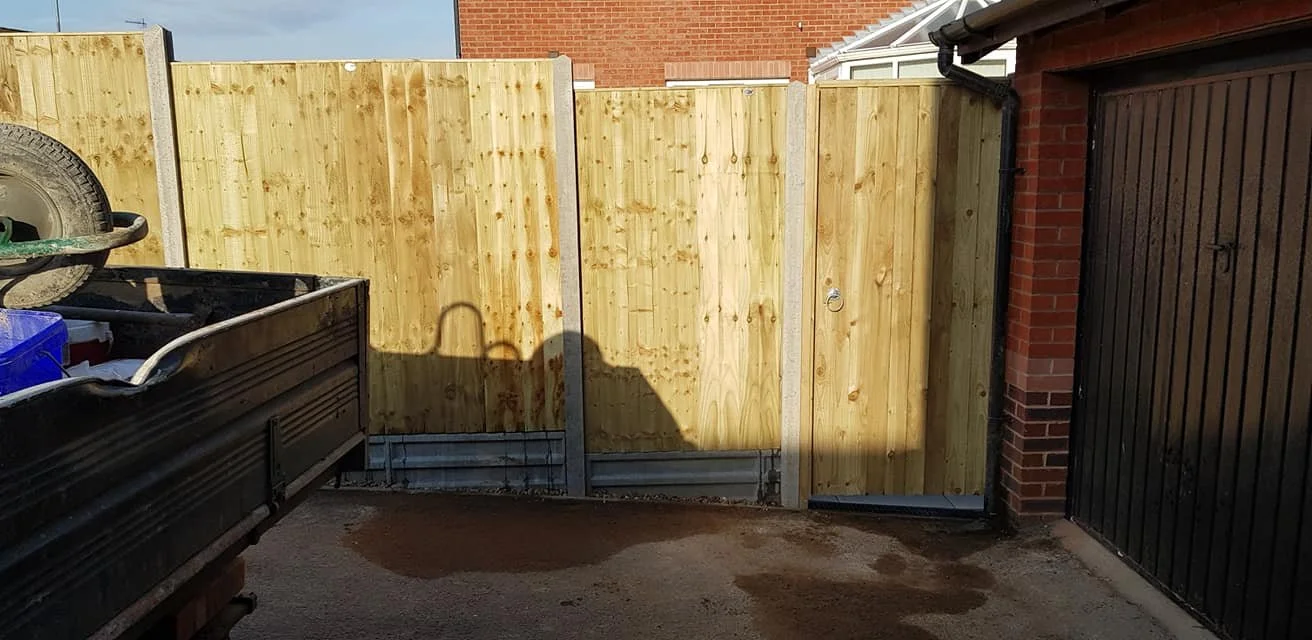View of a backyard construction area with a new wooden fence, a black gate, and a brick building wall.