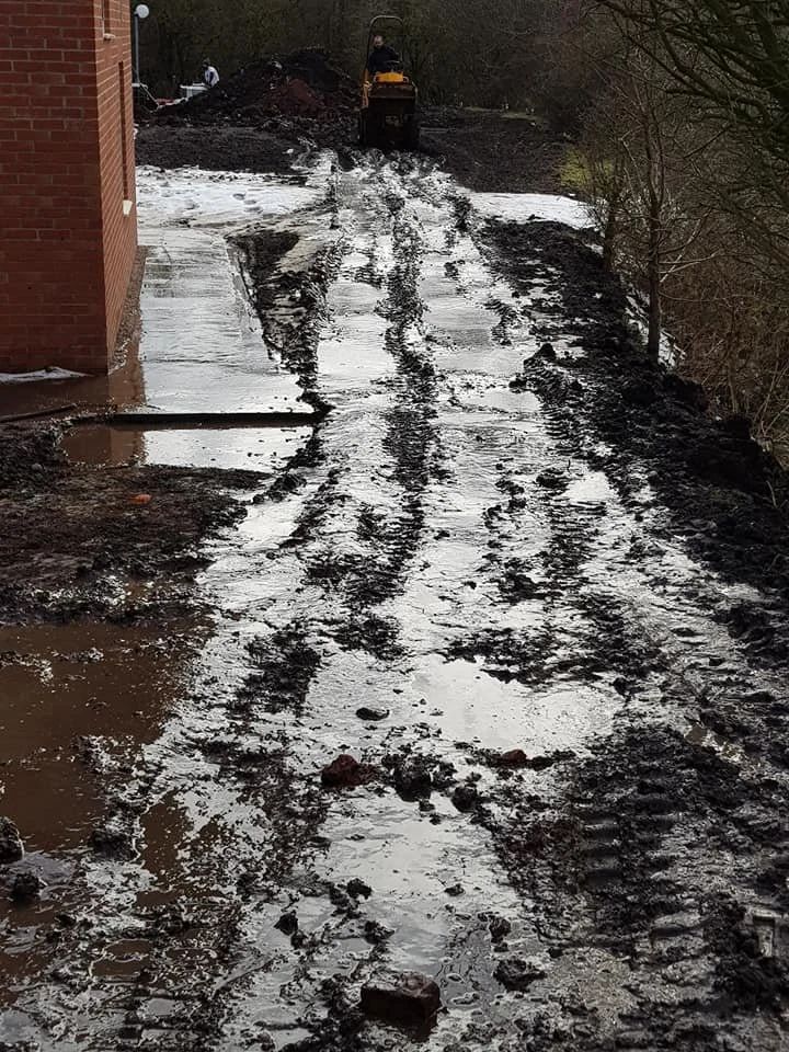 A muddy, partially frozen dirt road with tire tracks and puddles, showing signs of recent heavy rain or flooding. There is construction equipment and a worker visible in the background.