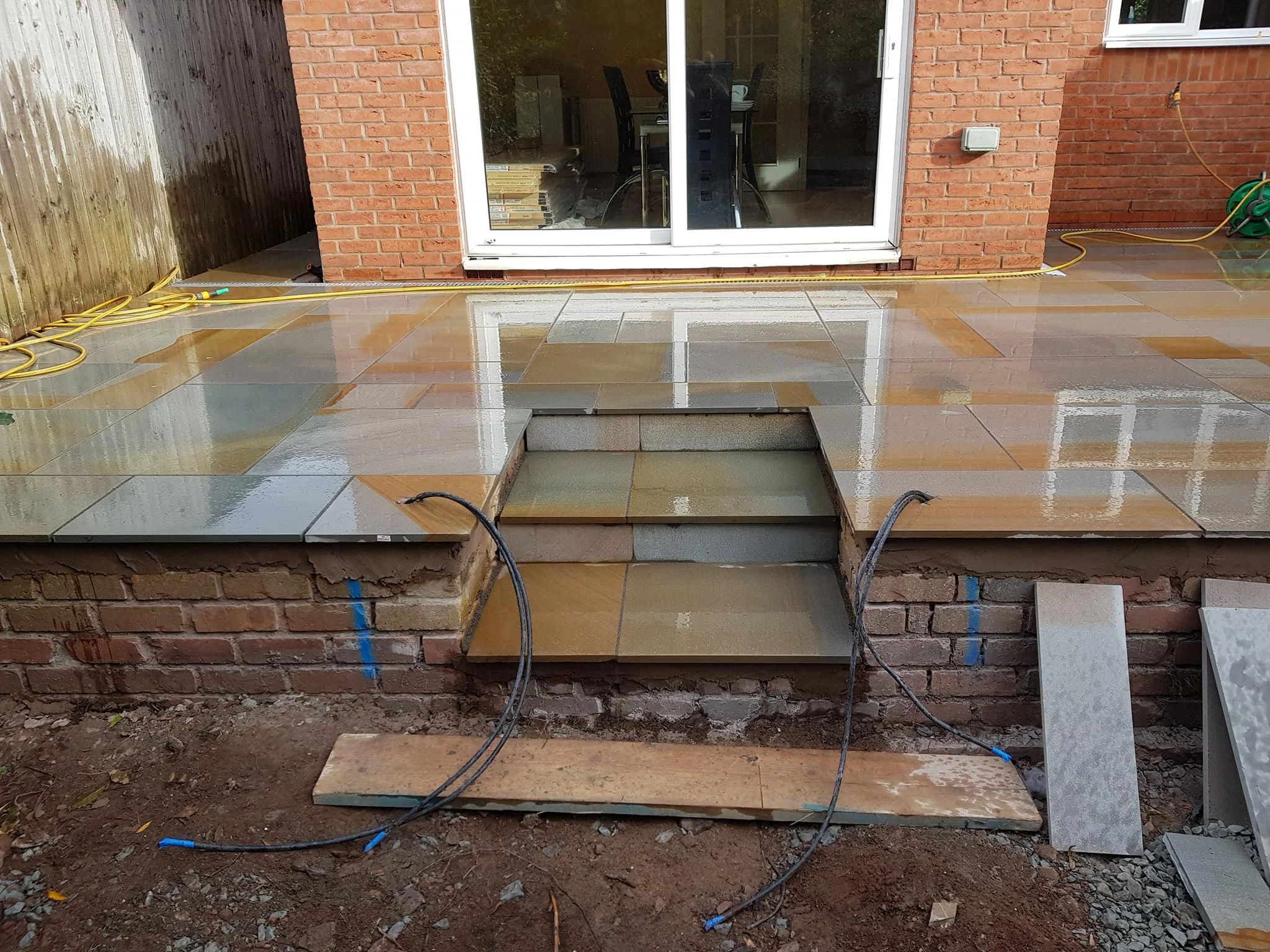 Construction of a brick and stone patio with steps in front of a glass sliding door on a brick house, with construction materials and electrical cords visible.