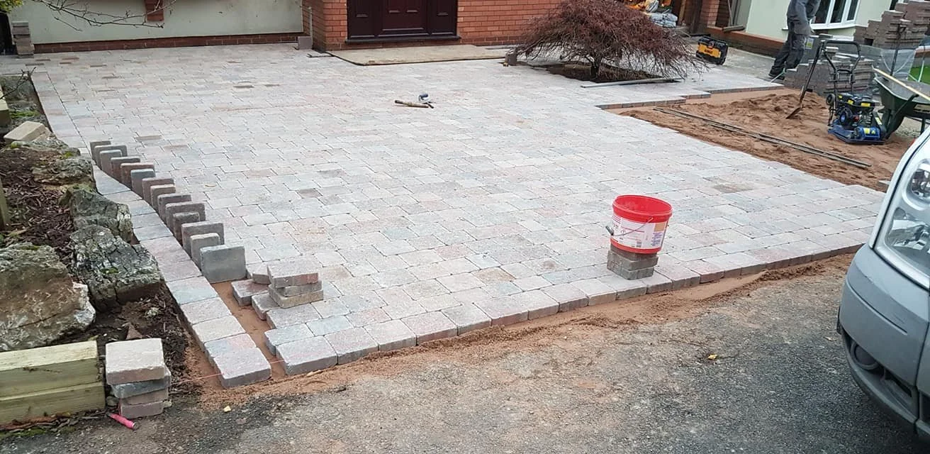 Paving stones are being installed to create a new driveway or patio area in front of a house, with some stones already laid and others waiting to be placed.