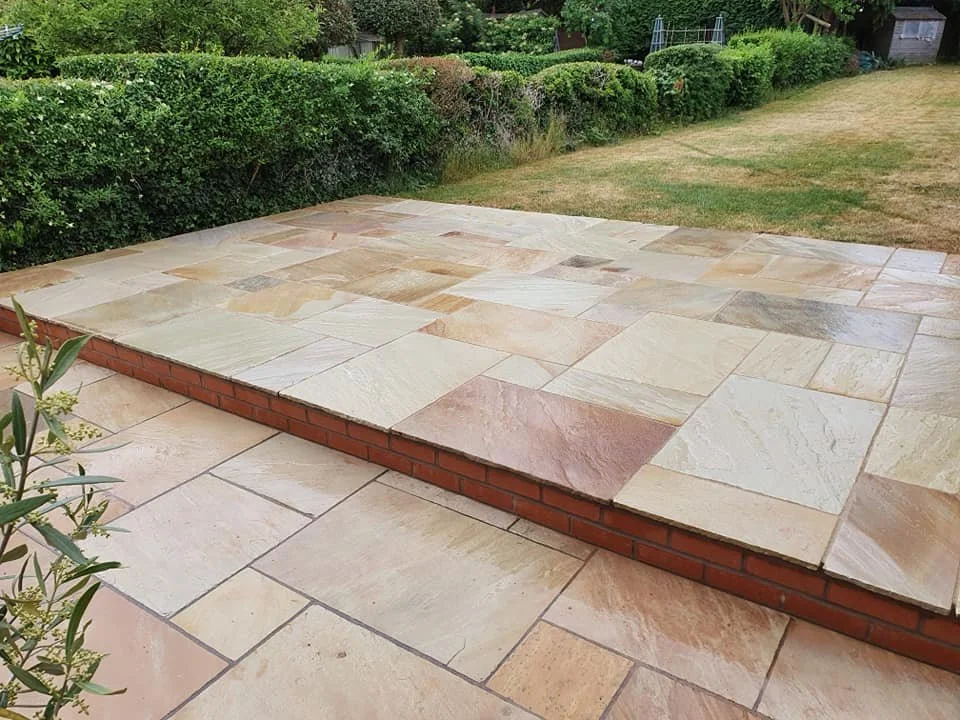 A backyard patio with a raised section made of light-colored stone pavers, bordered by a red brick edge, with a grassy lawn and green shrubbery in the background.
