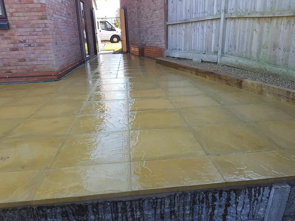 Wet yellow paving stones on a patio or walkway next to a brick wall and a wooden fence, with a white van visible through an open gate.