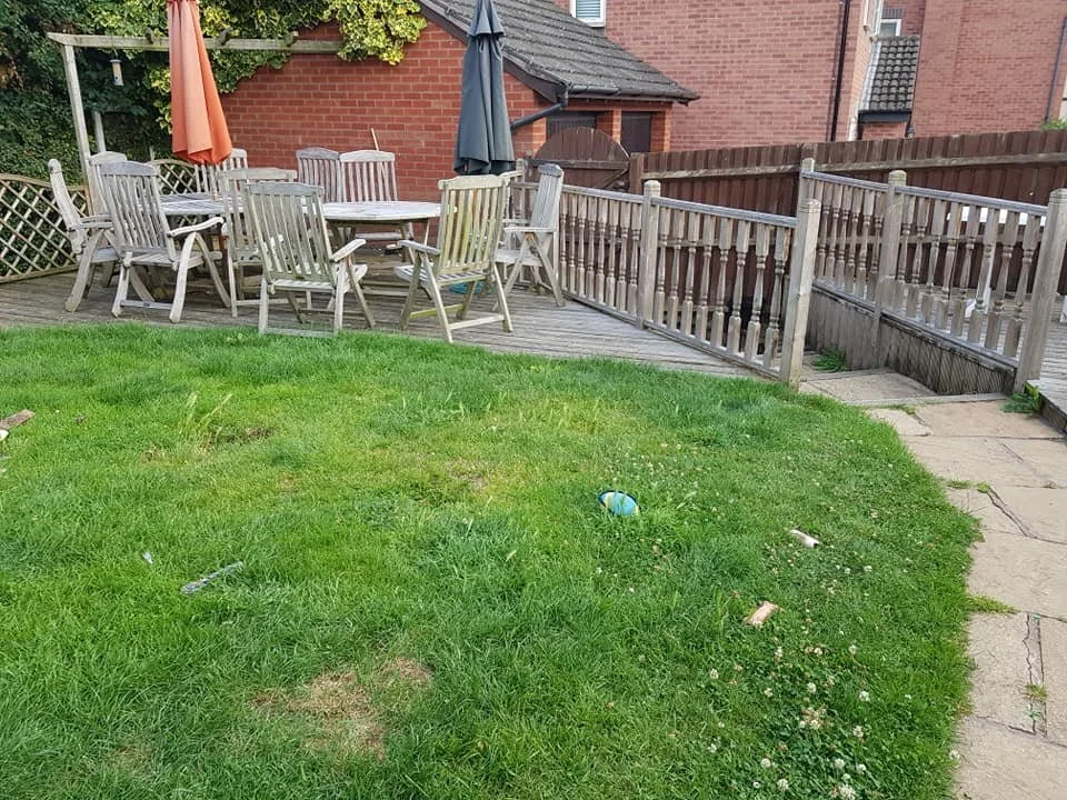 A backyard with a grassy lawn, a wooden deck with a round table and chairs, an umbrella, and a staircase leading down to a stone pathway. There are some small items and debris on the grass.