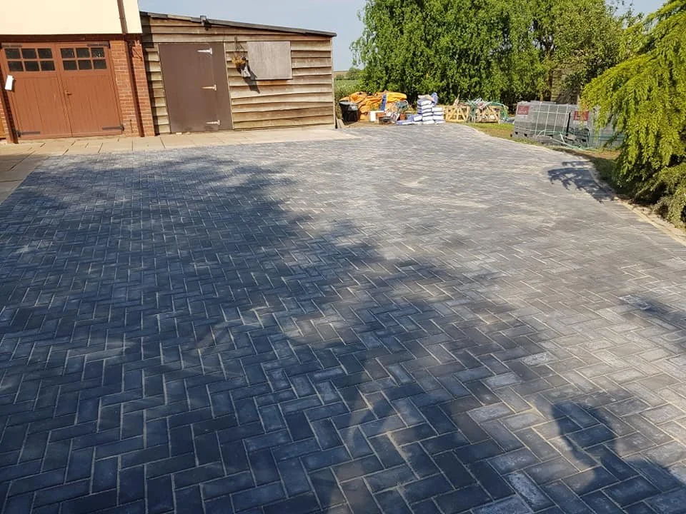 A newly paved driveway with dark gray bricks in front of a shed and a garage, with trees and outdoor storage in the background.