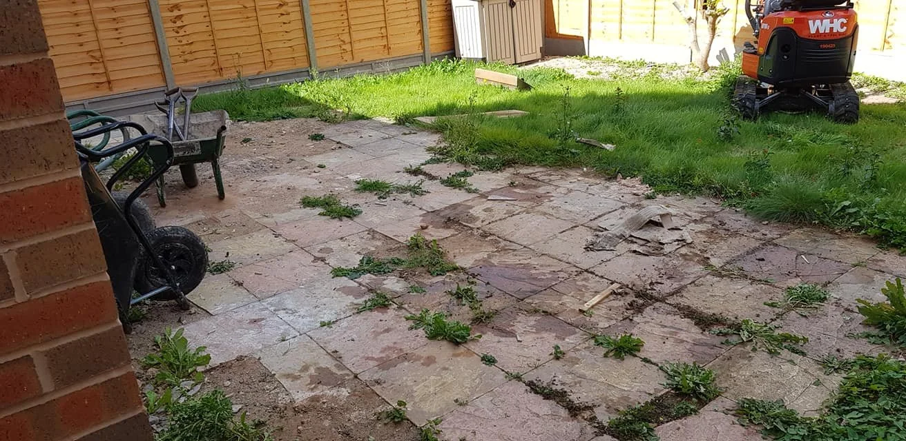Backyard patio with old uneven tiles, weeds growing between, a wheelbarrow, garden tools, a small red and black robot, and a wooden fence.