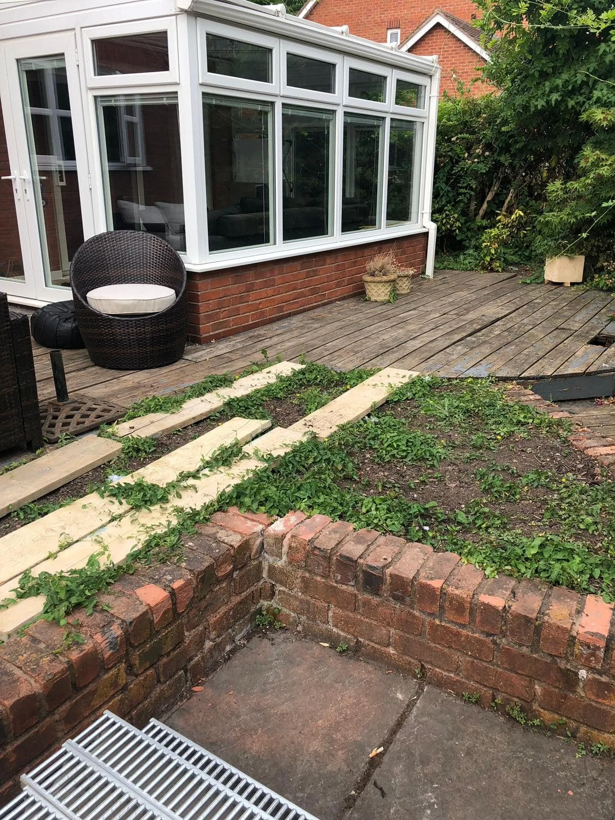 Backyard with a small wooden deck, a brick wall, a black wicker chair with a towel, and a greenhouse with glass windows attached to a brick house. There are plants and greenery surrounding the area.