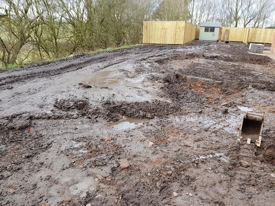Construction site with muddy ground, partially excavated yard, and a wooden fence in background.