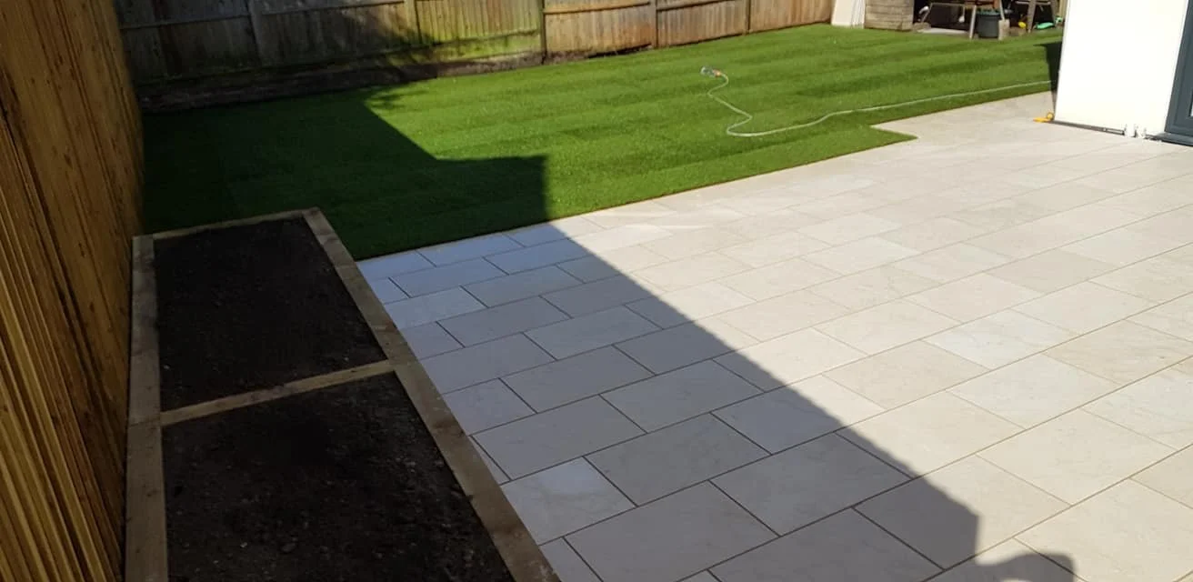 Backyard with newly laid white stone pavers and a patch of artificial grass. A garden bed is outlined with wooden framing, and the backyard is enclosed with a wooden fence.