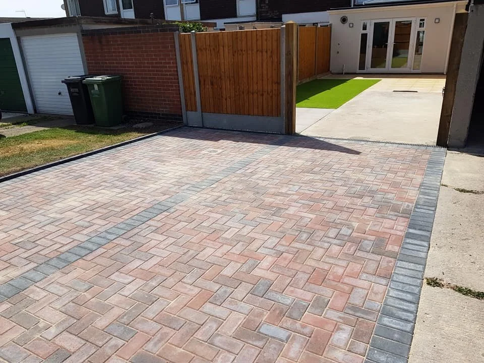 A paved driveway with interlocking bricks leading to a backyard with a concrete patio, a small strip of artificial grass, and a wooden fence.