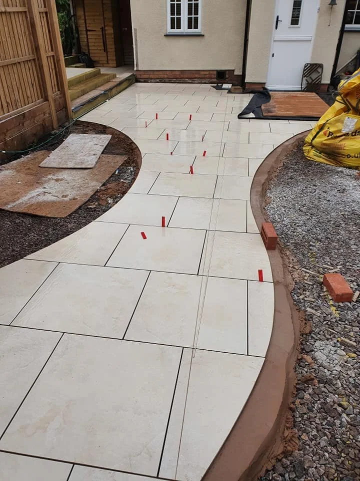 Installing new beige ceramic tiles on a curved outdoor patio with orange spacers and construction materials on the ground.