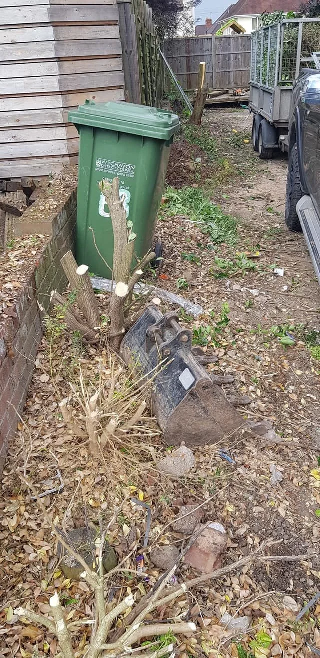 A garden area with a green trash bin, a tree stump with pruned branches, a black tractor bucket attachment, and a parked vehicle on the right, surrounded by leaves, small plants, and a wooden fence.