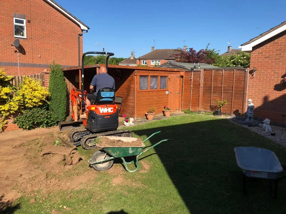 A person operating a mini excavator in a backyard, with a wheelbarrow and various garden decorations, including potted plants, on a sunny day.