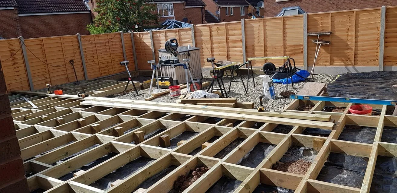 Construction site for a wooden deck with a grid of wooden beams, surrounded by a tall wooden fence, with tools and materials scattered around.