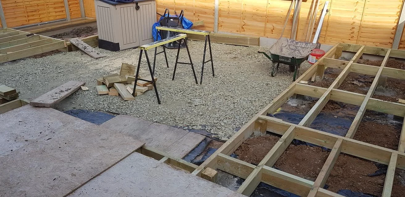 Construction site with wooden framework, gravel ground, tools, and workbench inside a fenced yard.