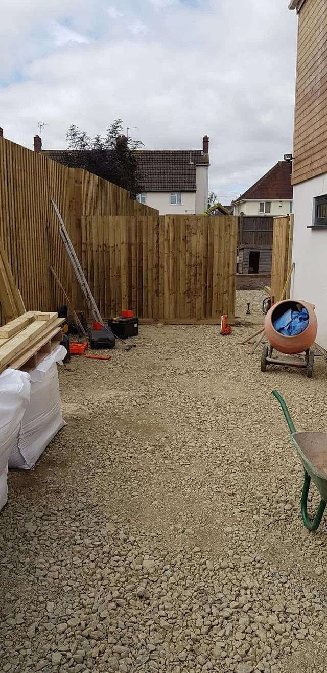 A backyard construction site with a gravel ground, wooden fencing, construction tools, a wheelbarrow, and a concrete mixer.