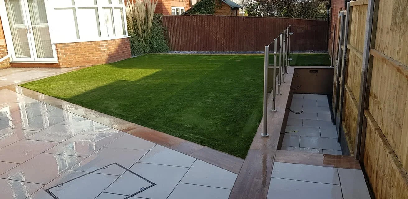Backyard patio with wet, shiny white tiles, a grassy lawn, a fenced area, and a small staircase with a metal railing leading to a lower level.