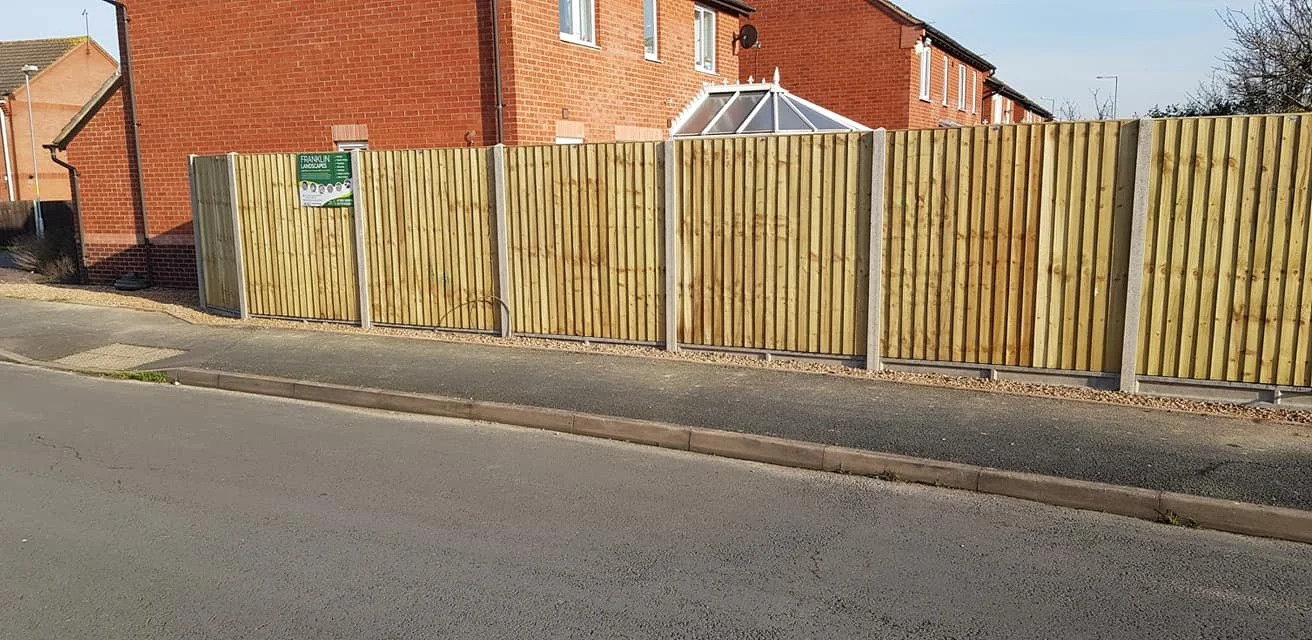 Wooden privacy fence along a sidewalk in front of a red brick residential building with windows and a sunroom extension.