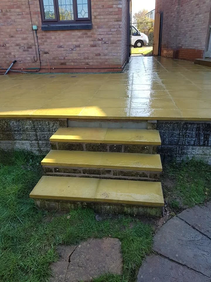 Bright yellow painted concrete stairs leading from grassy area to a raised concrete patio at the back of a brick house, with a window and a door opening to the patio and a view of a white vehicle parked outside.