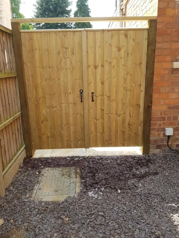 Backyard wooden gate with two black handles, brick wall on the right, wooden fence on the left, and a concrete slab in the foreground, surrounded by gravel.