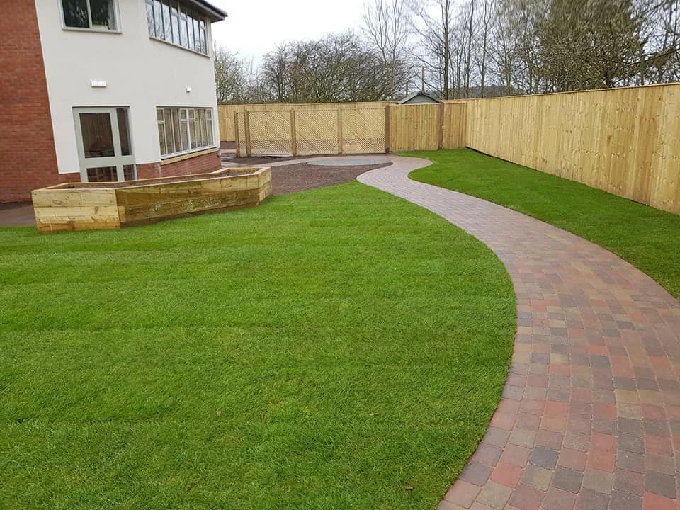 A backyard with a curved brick pathway, green lawn, wooden fence, and a portion of a house with large windows.
