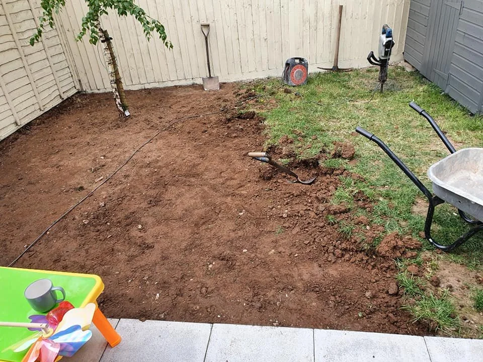 A backyard garden area with soil prepared for planting, surrounded by grass and a fence, with gardening tools, a wheelbarrow, and a tree in the corner.