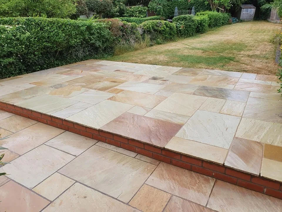 A backyard patio with beige and light pink stone tiles, a brick border, and a large grassy area with trees and shrubs in the background.