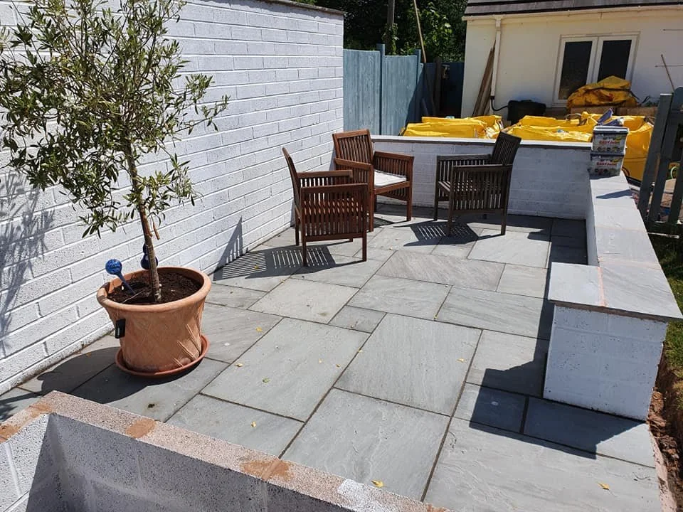 A newly constructed outdoor patio with light gray slate tiles, a white brick wall on the left, a potted olive tree, and garden furniture including four wooden chairs. In the background, there are construction supplies covered with yellow tarps and a 