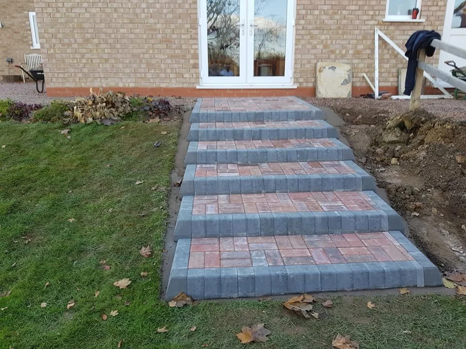 Completed brick and concrete stairway leading up to a house with a brick exterior and large glass doors, with some construction debris and soil on the sides.