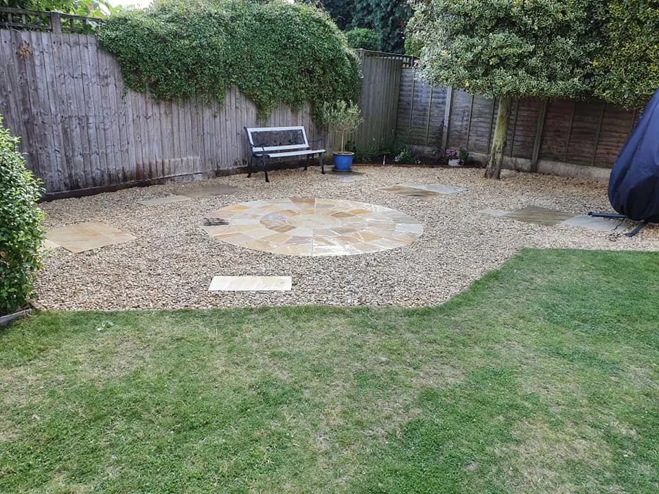 A backyard with a small grassy lawn, a wooden fence, and a gravel patio area with a circular pattern of stone tiles. There's a black and white bench, a blue potted plant, and some trees and bushes.