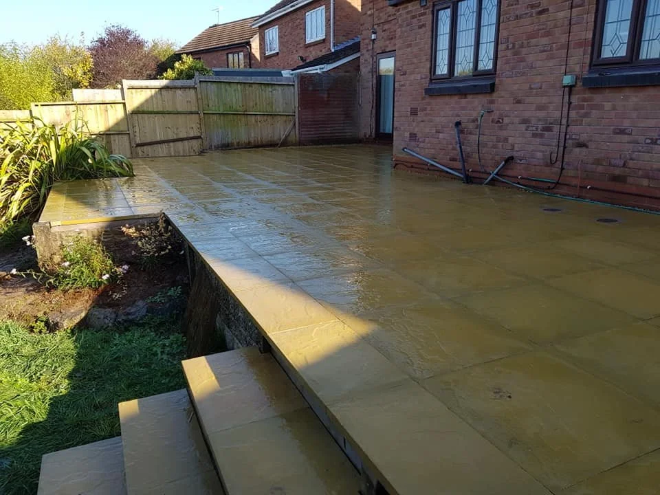 A newly paved backyard patio with beige stone tiles, adjacent to a brick house with three windows, and a green garden fence in the background.