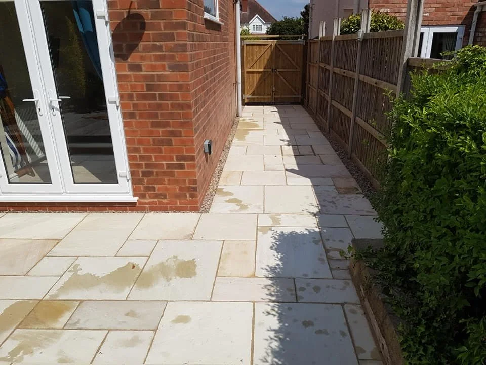 Backyard patio with beige stone tiles, wet from recent cleaning, next to a red brick house with white double doors, enclosed by a wooden fence with a wooden gate at the end, and green bushes on the right side.