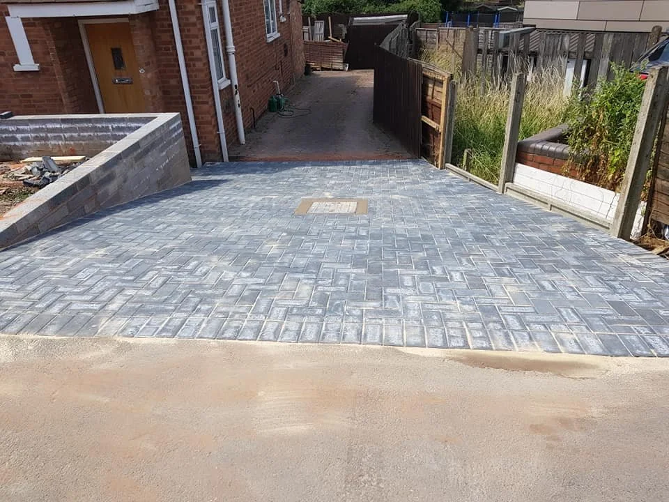 A newly paved driveway with gray bricks leading to a back alley, with a brick house on the left and a wooden fence on the right.
