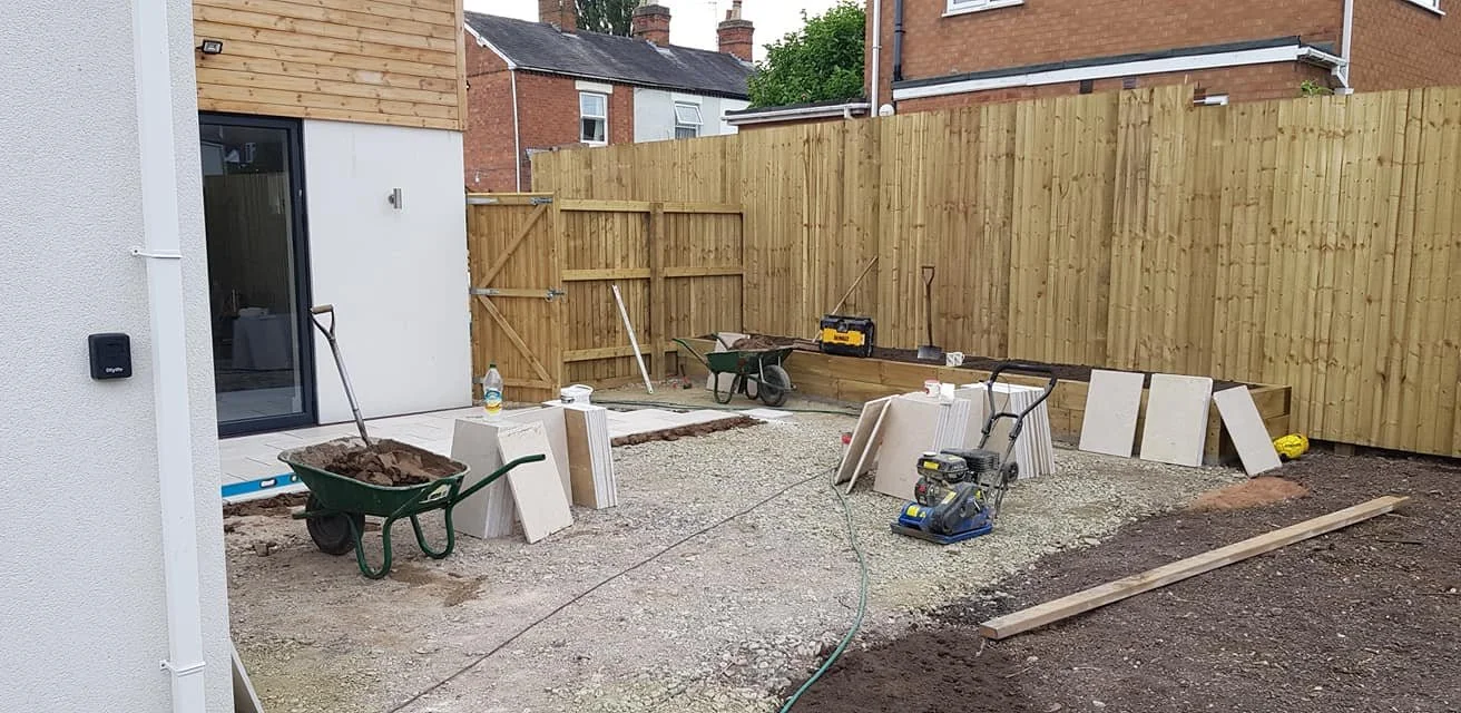 Backyard under construction with wheelbarrows, pavers, and tools. New wooden fence being built around the yard.
