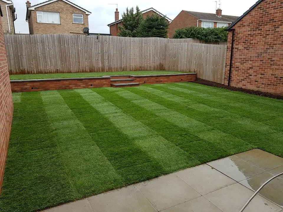 A backyard with newly laid striped green grass, a brick retaining wall with steps, surrounded by wooden fencing and neighboring houses in the background.