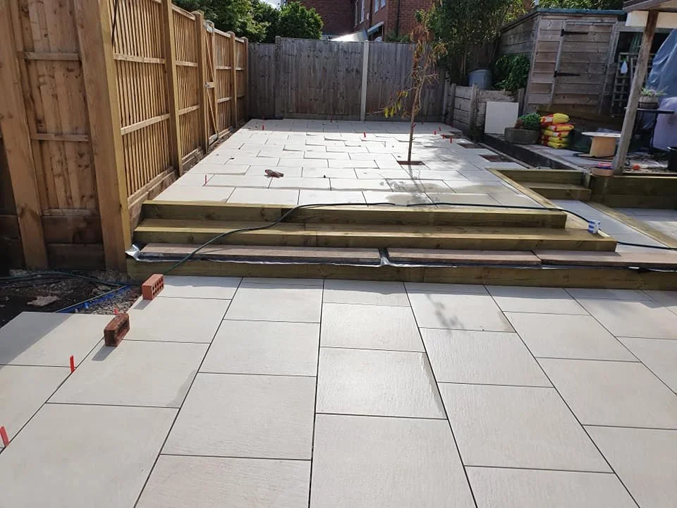 A backyard in the process of paving, with new concrete tiles laid and wooden steps being constructed, surrounded by a fence, with some garden supplies and tools nearby.