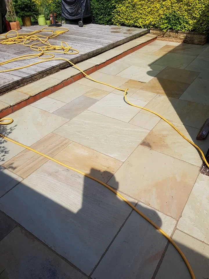 A backyard patio with freshly laid beige and light brown stones, a yellow extension cord running across the stones, and a wooden deck with some plants and gardening tools in the background.