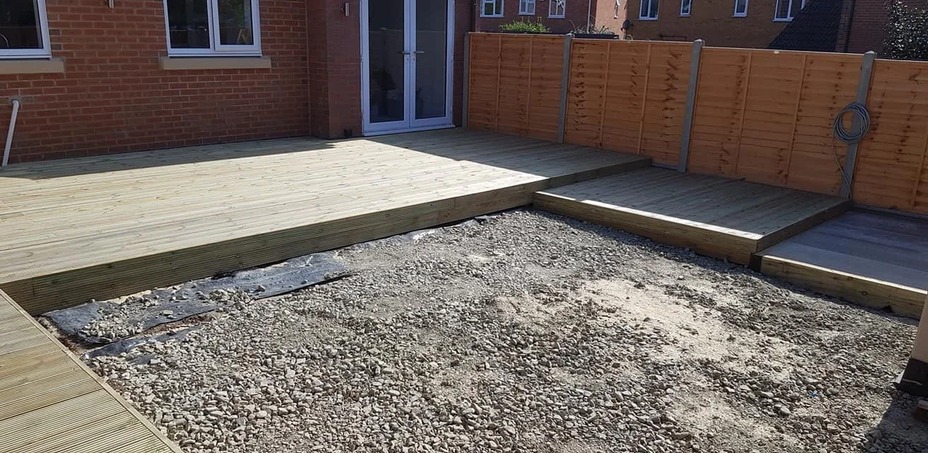 A backyard with a new wooden deck partially built, with gravel and dirt ground, and a wooden fence around the yard.