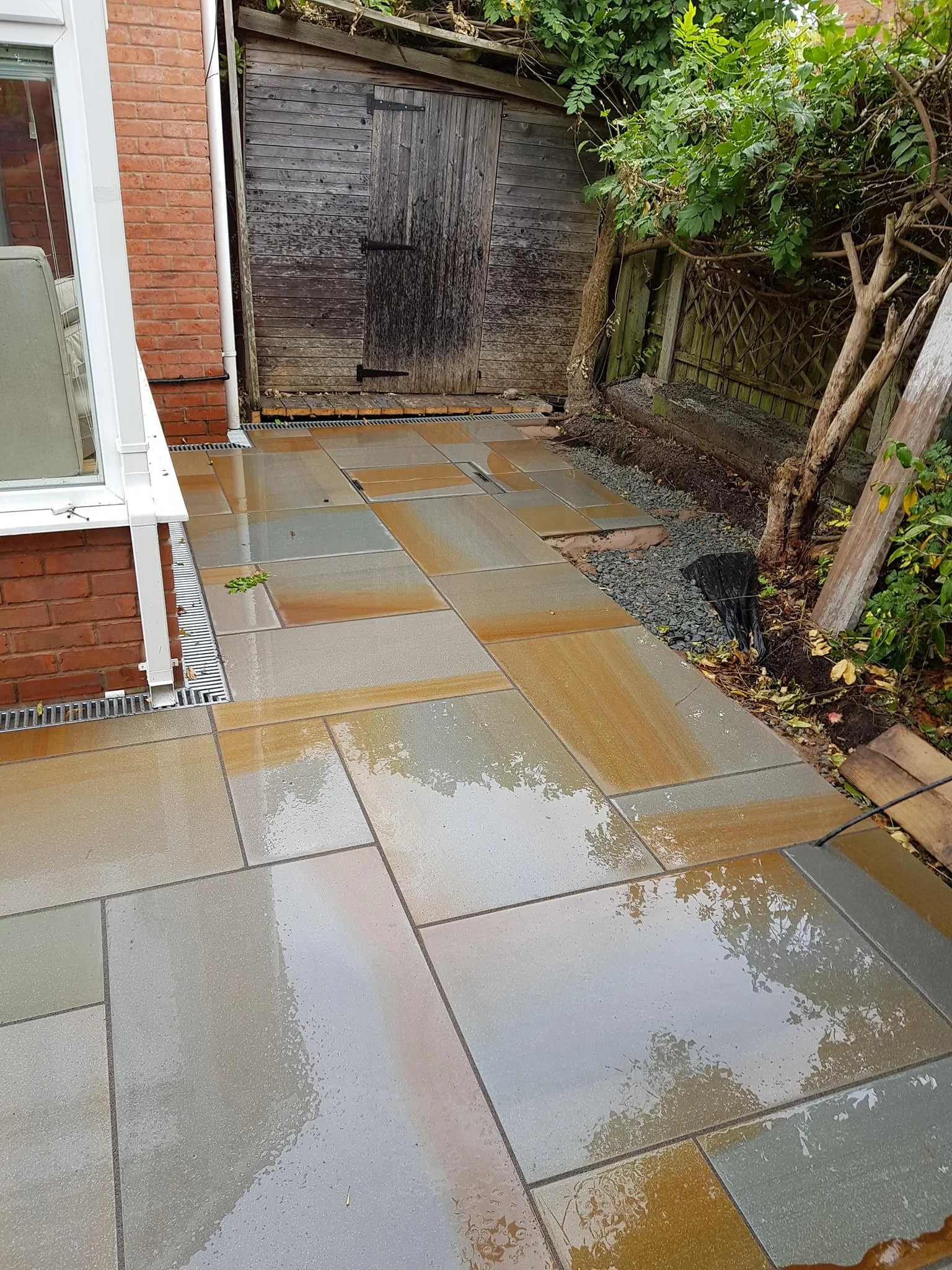 Wet outdoor patio with multicolored stone tiles, bordered by a red brick wall, a wooden shed in the background, and greenery on the right side.